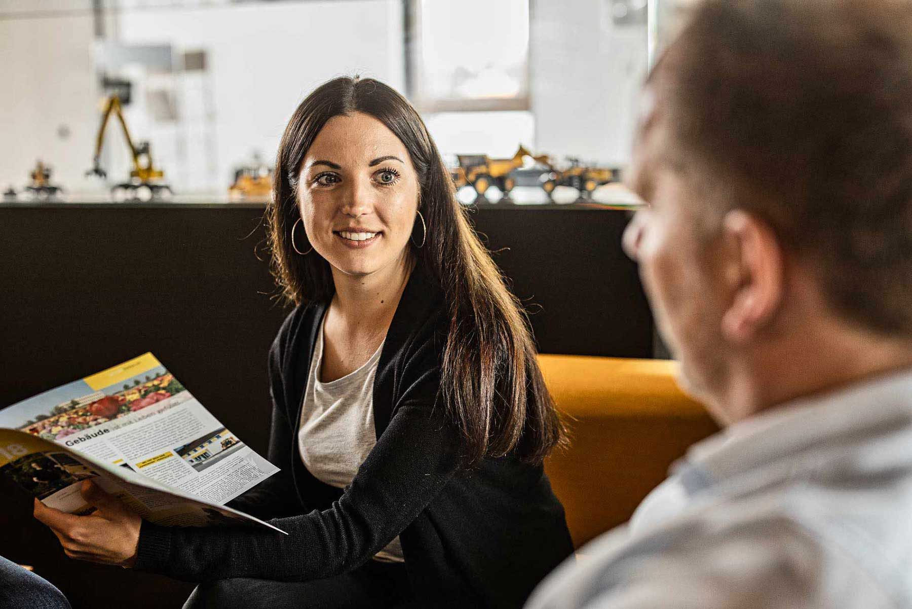 Unternehmensfotografie von einer frau die einem Mann ein Magazin zeigt. Sie sitzen beide auf einem gelben Sofa.