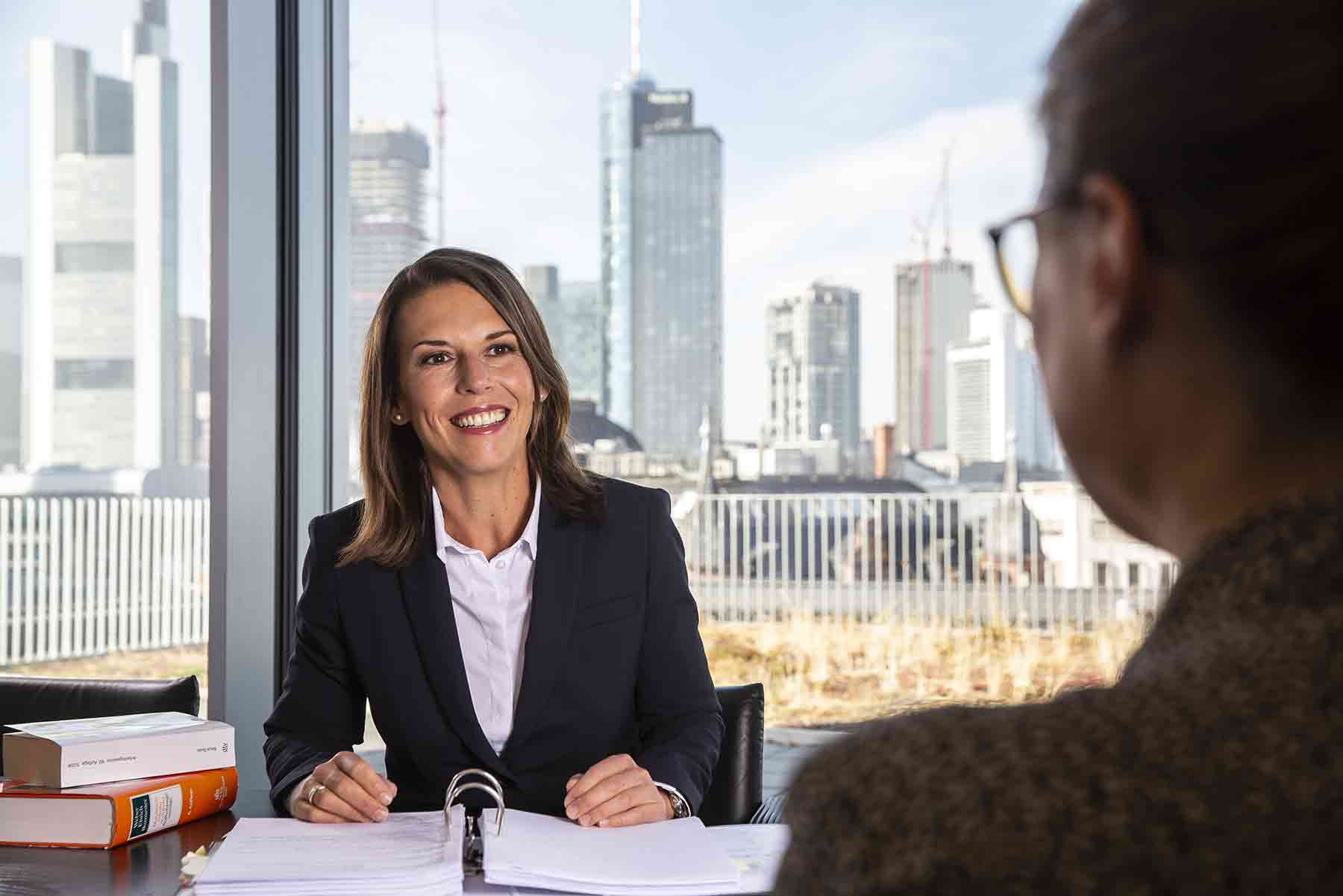 Unternehmensfotografie und Businessportrait einer Frau am Schreibtisch. Im Hintergrund ist eine Stadt zu sehen.