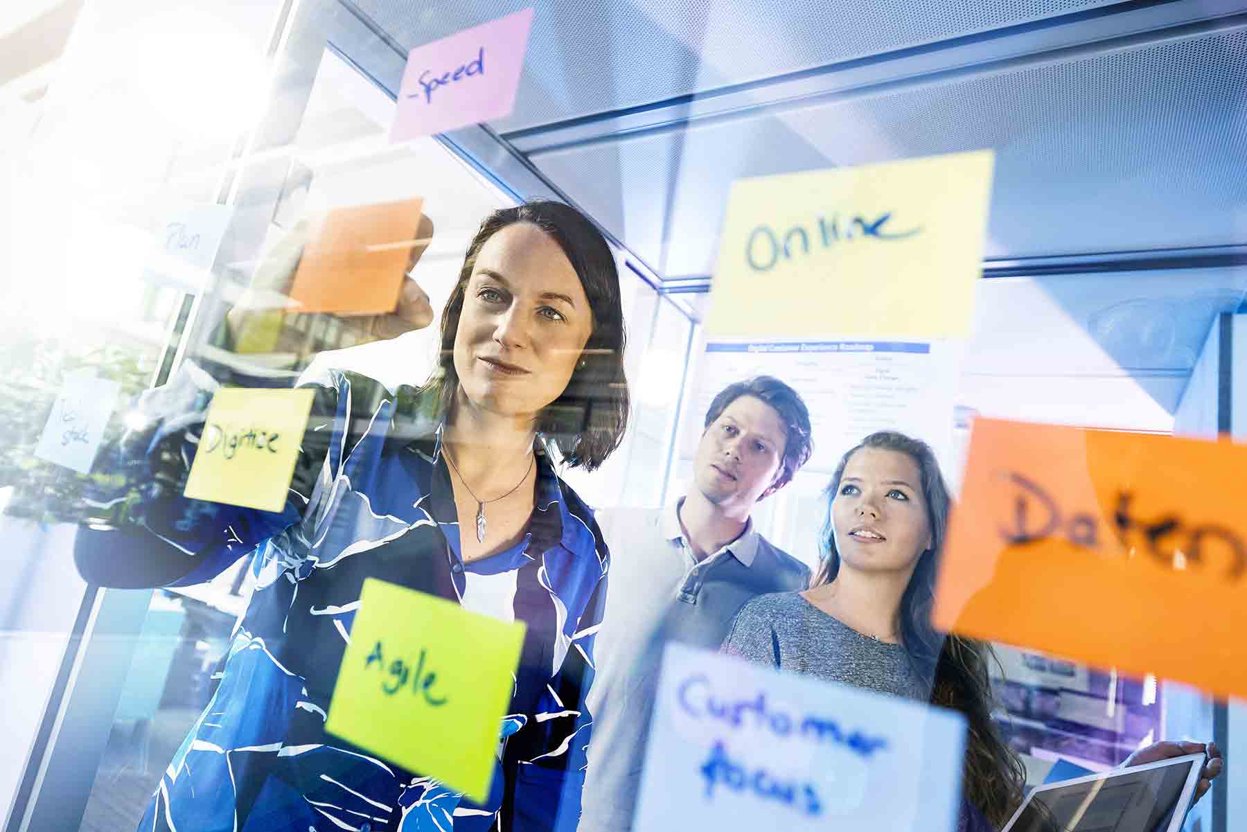 Ein Businessportrait einer Frau, die auf bunte Blätter schreibt. Zwei Personen im Hintergrund