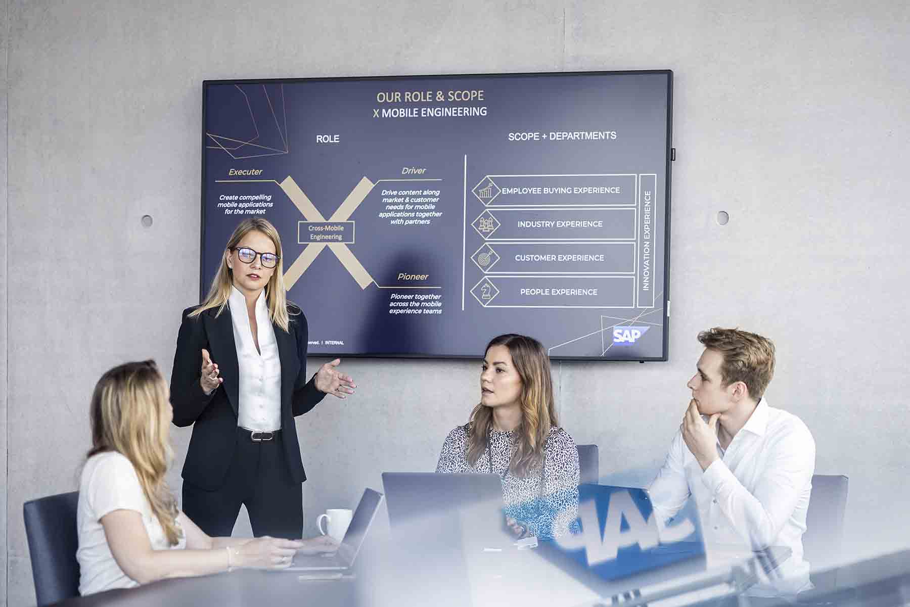 Businessportrait einer Frau, die vor anderen referiert. Im Hintergrund ist ein großer Monitor.Im Vordergrund spiegelt sich der Schriftzug SAP wieder.