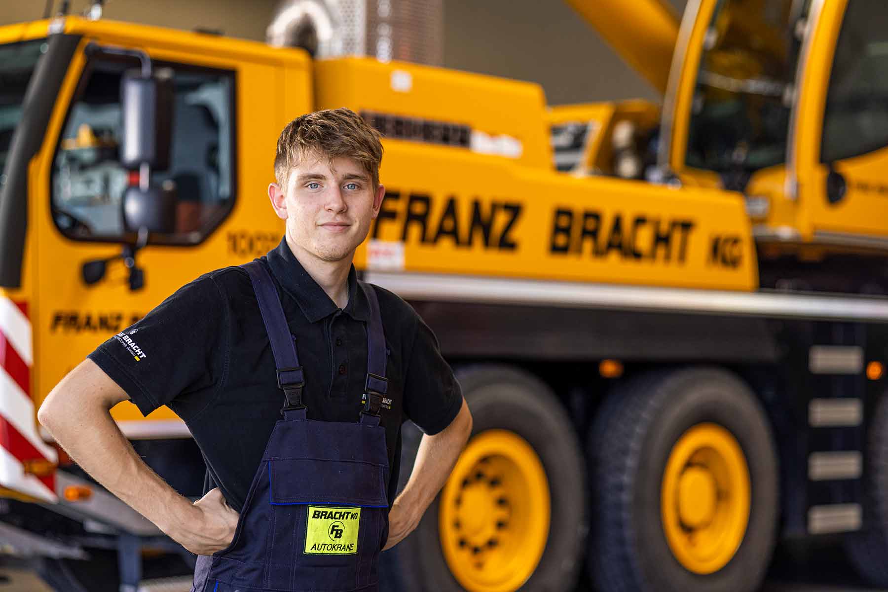 Ein junger Azubi im Blaumann steht vor einem Baufahrzeug.